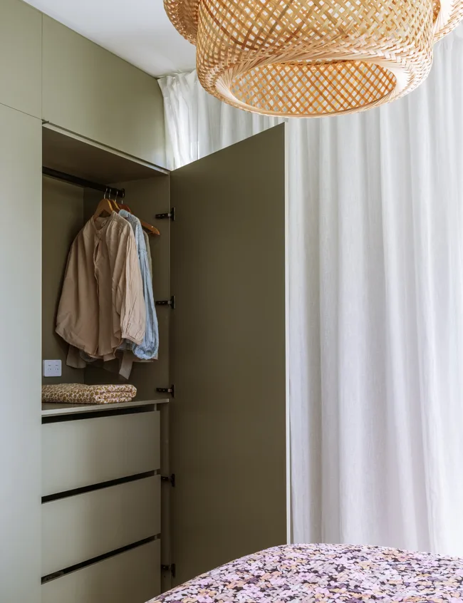 Wardrobe with open door showing hanging clothes and folded sheets; white curtains and woven ceiling light above.