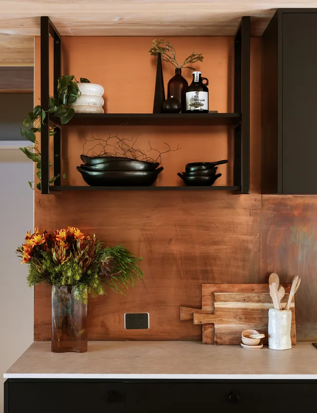 Rustic kitchen shelf with plants, dark dishes, wooden boards, and a vase of colorful flowers on a countertop.