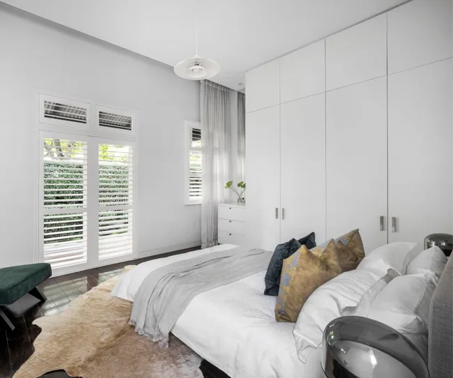 Modern bedroom with white walls, a bed with decorative pillows, and large windows with plantation shutters.