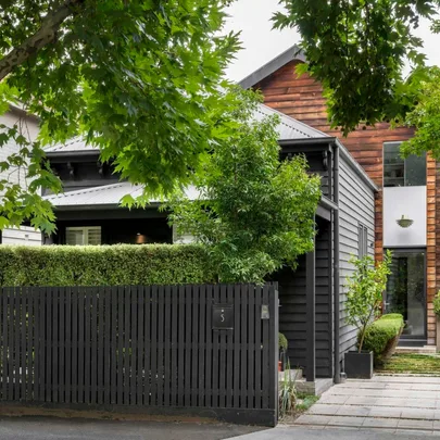 Modern two-story house with dark wood siding, surrounded by trees and greenery, and a black picket fence in front.