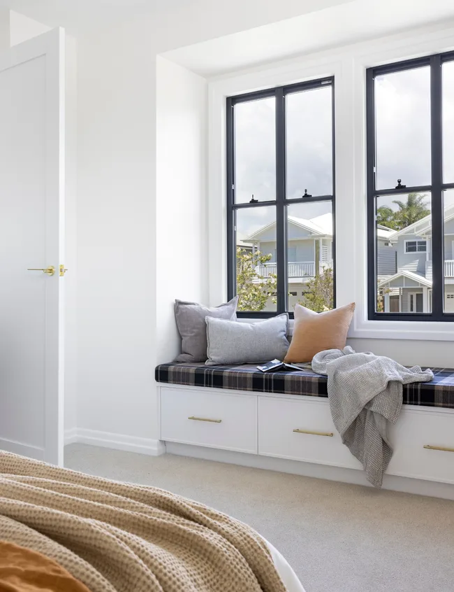 Cozy window seat with cushions and blanket in a bright room, overlooking houses through large windows.