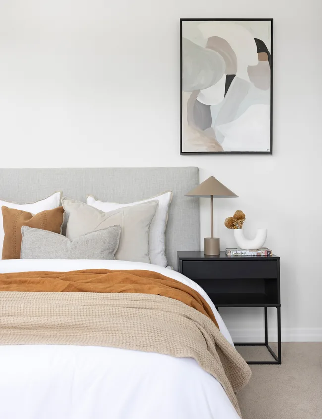 Modern bedroom with gray headboard, abstract art, beige and white bedding, tan throw, and black nightstand with lamp.