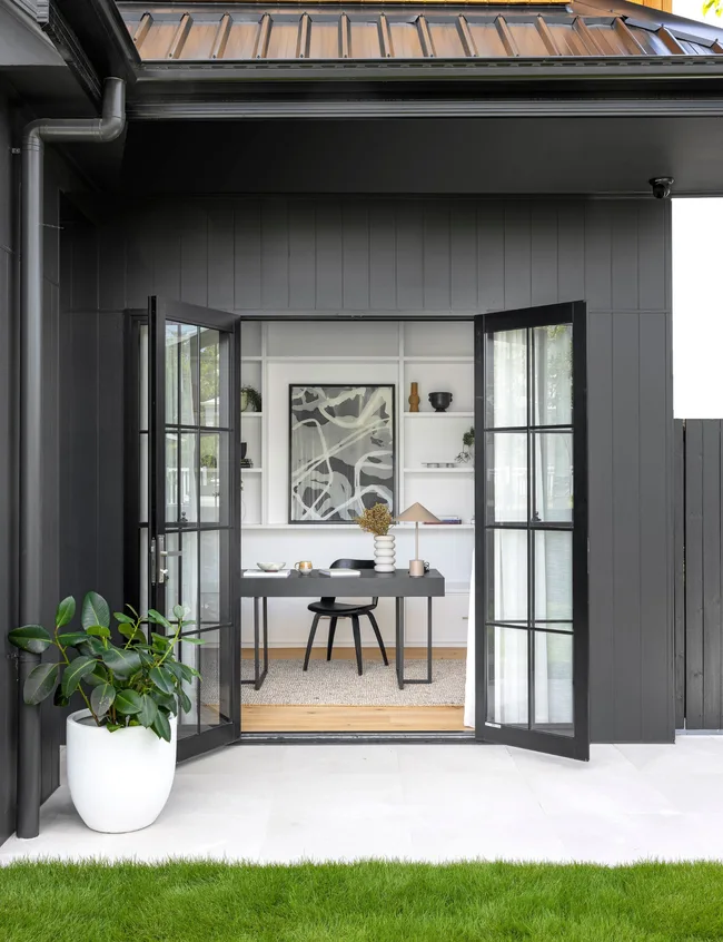 Modern home office with open black-framed doors, black desk, abstract art, and white shelves; plant on patio.