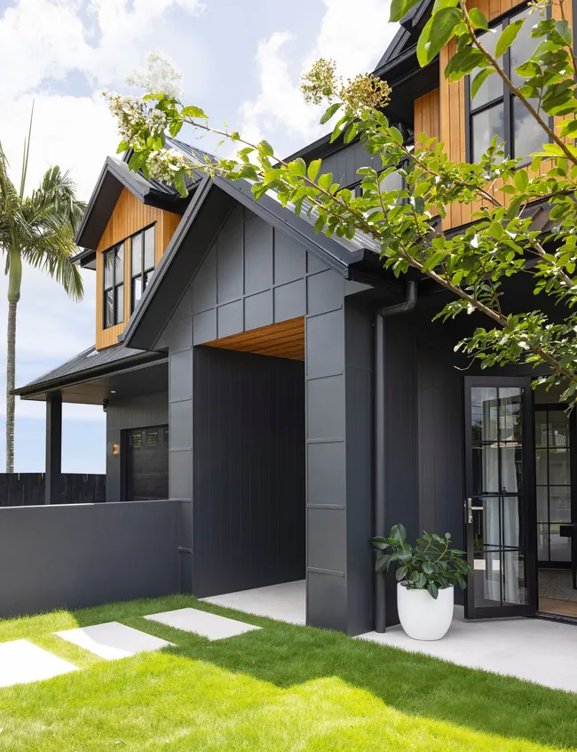 Exterior of a modern house with black paneling, wood accents, and lush greenery, featuring a small garden path.