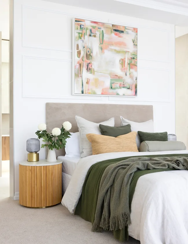 Modern bedroom with abstract art, cozy bedding in green tones, wooden side table, and decorative pillows and flowers.