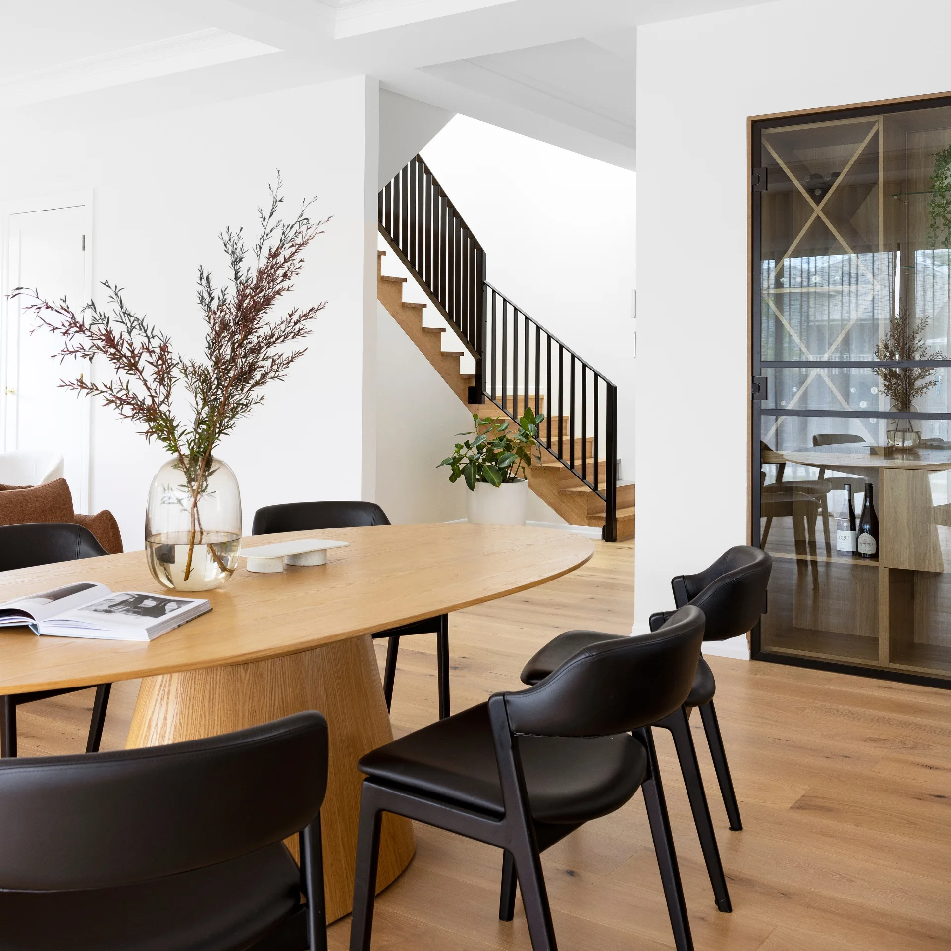 Modern dining area with wooden table, black chairs, staircase, and a plant. A decorative vase with branches sits on the table.
