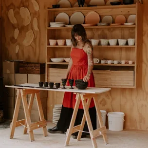 Woman in a red dress arranging ceramics on a table in a pottery studio with shelves of stacked bowls behind her.