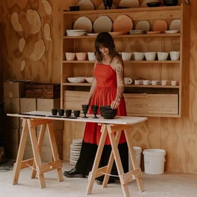 Woman in a red dress arranging ceramics on a table in a pottery studio with shelves of stacked bowls behind her.