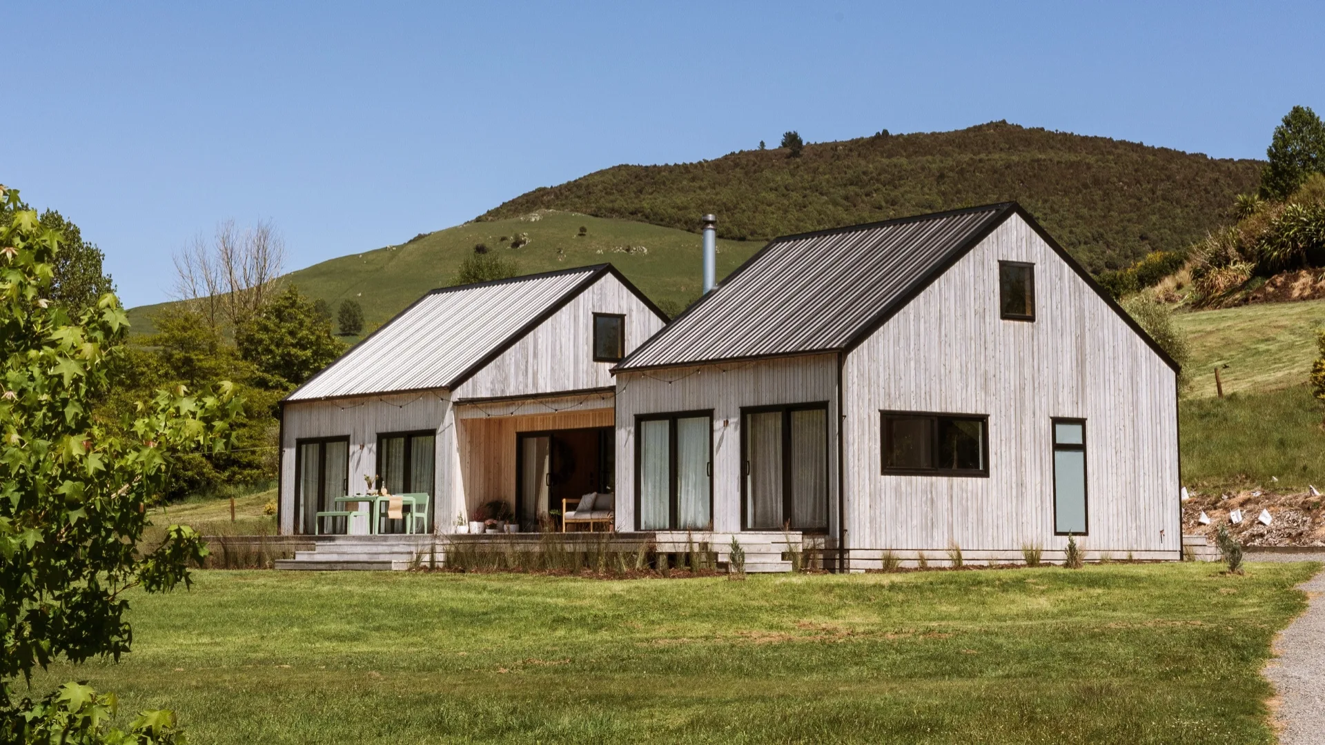 Two rustic wooden houses with metal roofs surrounded by lush greenery and hills under a clear blue sky.