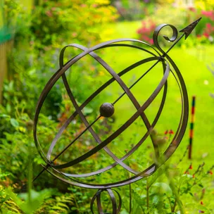 Armillary sphere sculpture in a vibrant garden setting with green grass and colorful flowers in the background.