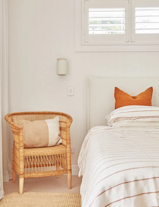 Cozy bedroom with a wicker chair, beige cushion, and white bed with orange pillow, under a window with shutters.