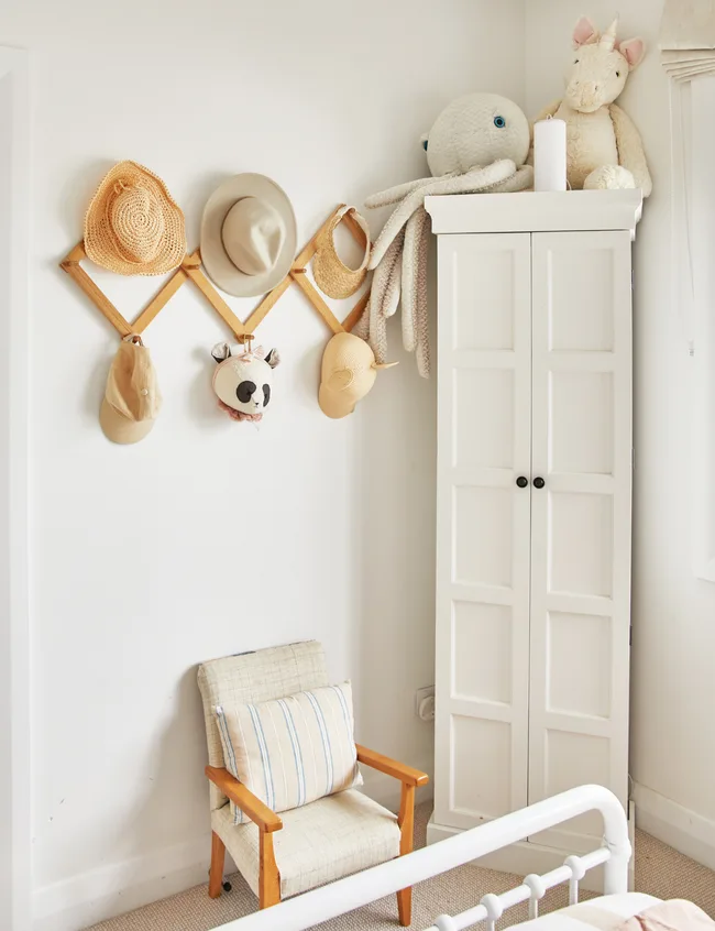 A cozy kid's room corner with hats on hooks, plush toys on a wardrobe, and a small chair with a cushion.