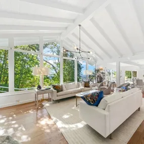 Bright living room with white sofas, high ceiling, modern chandelier, large windows, and forest view.