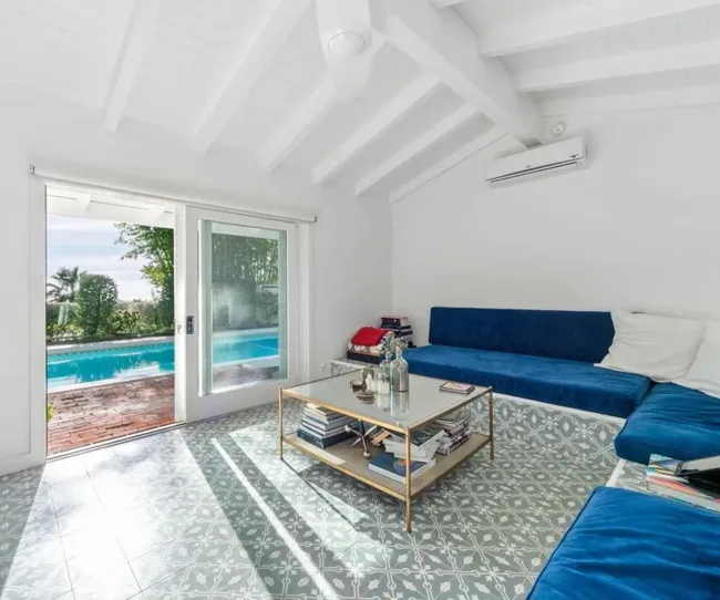 Bright living room with blue sofas, patterned tile, glass coffee table, and sliding door leading to pool and garden.