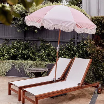 Two wooden lounge chairs with white cushions under a pastel pink fringed umbrella in a garden setting.