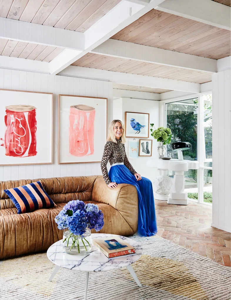 A woman in a blue skirt and a leopard print top sitting on the arm of a leather sofa with coco-cola art behind her.