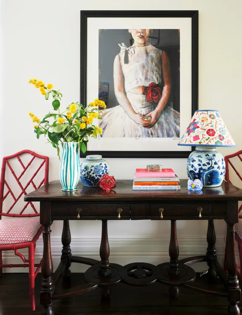 A vase of colourful flowers in a vase on a console table with books and a bright, multi-coloured lamp.