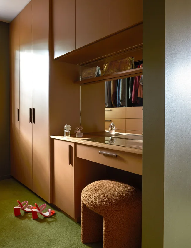 Brown closet and vanity with perfume bottles, brown stool, red high heels on green carpet.