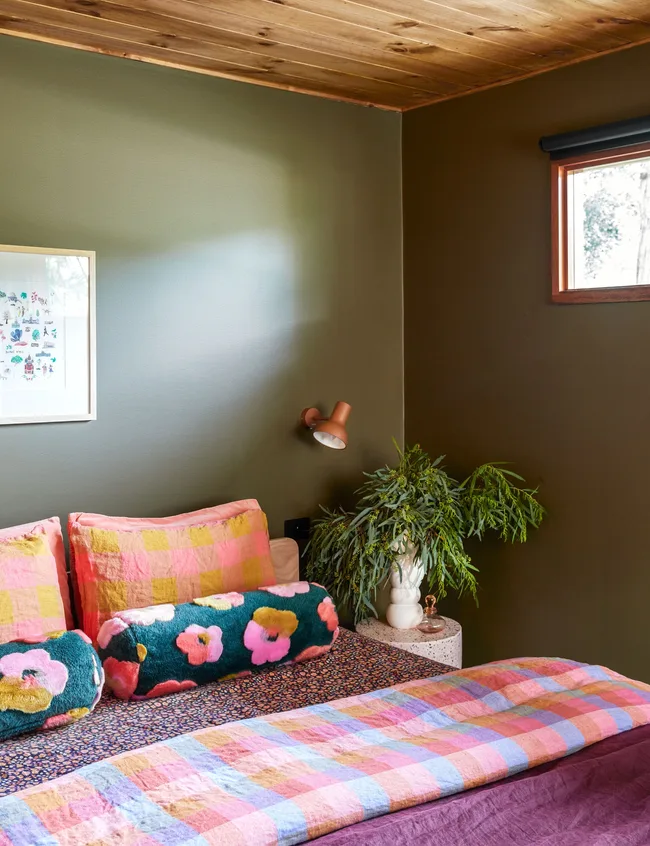 Cozy bedroom with colorful bedding, floral cushions, green wall, and wooden ceiling. Plant and framed art add decor.