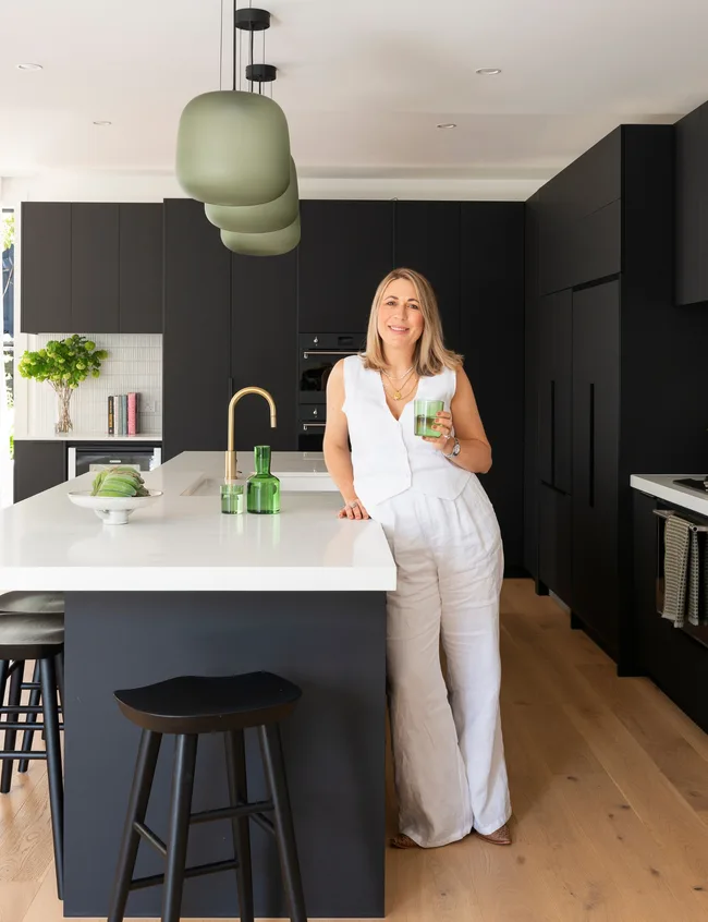 Woman in a modern kitchen with black cabinets, white countertop, holding a green glass, under green pendant lights.