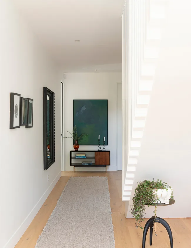 Modern hallway with light wood floor, framed art, green painting, small table with plant, and white staircase.