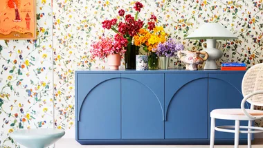 Vases of bright flowers sit on a blue sideboard with floral wallpaper behind.