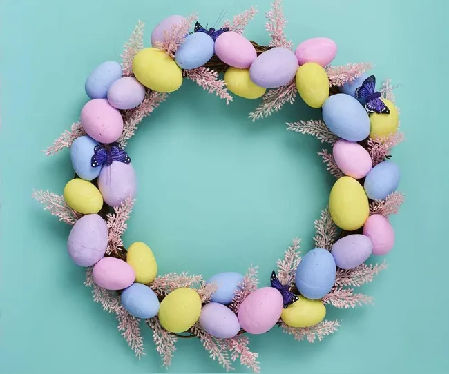 Wreath made of pastel-colored eggs and pink branches with blue butterflies on a teal background.