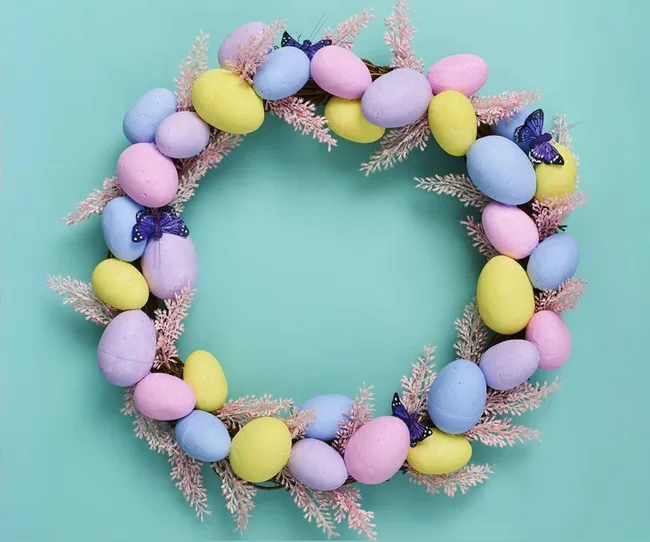 Wreath made of pastel-colored eggs and pink branches with blue butterflies on a teal background.