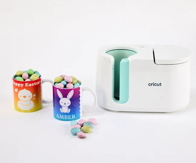 Cricut machine with two Easter-themed mugs filled with colorful candy-coated eggs; one mug has "Amber" written on it.