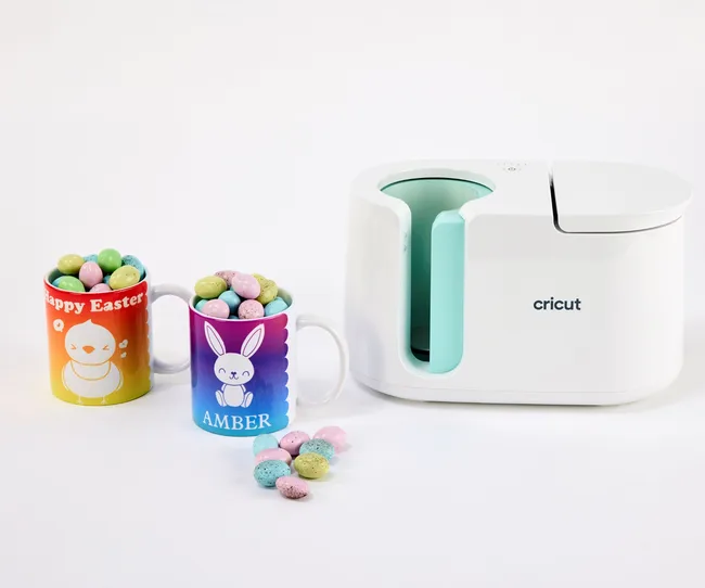 Cricut machine with two Easter-themed mugs filled with colorful candy-coated eggs; one mug has "Amber" written on it.