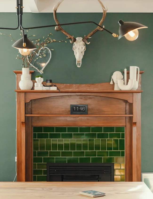 Wooden fireplace with green tiles, antler skull above, vase, and decor items on mantelpiece; wall-mounted clock shows 11:46.