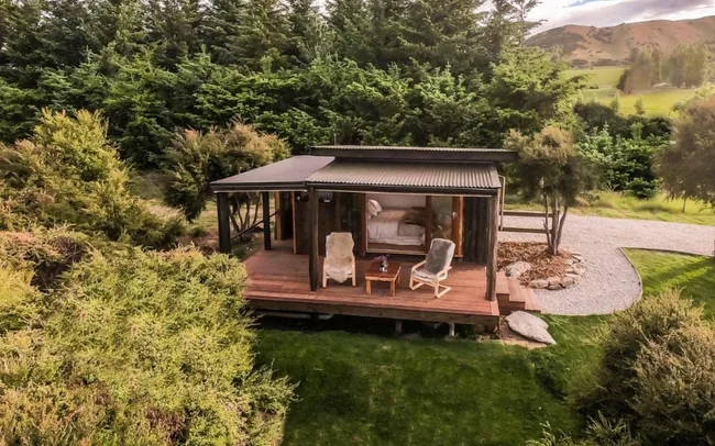 Rustic cabin with a deck, surrounded by greenery and distant hills under a cloudy sky.