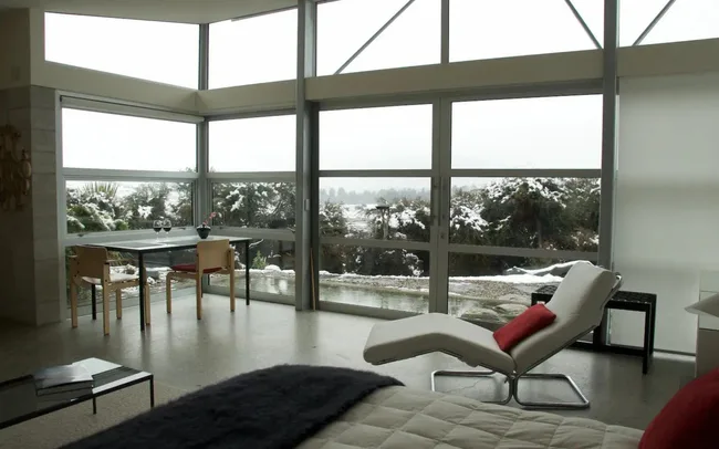 Modern living room with large windows, white chaise lounge, table with chairs, and snowy outdoor view.