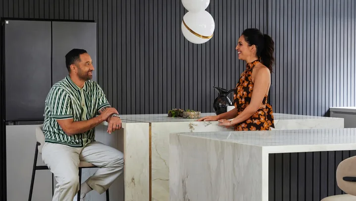 Two people converse in a modern kitchen with marble countertops and a stylish pendant light.