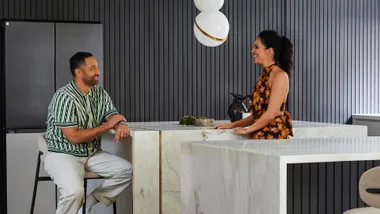 Two people converse in a modern kitchen with marble countertops and a stylish pendant light.