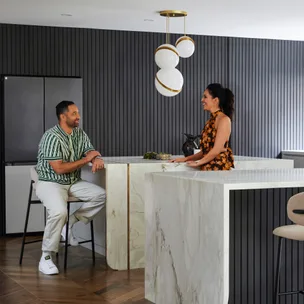 Two people converse in a modern kitchen with marble countertops and a stylish pendant light.