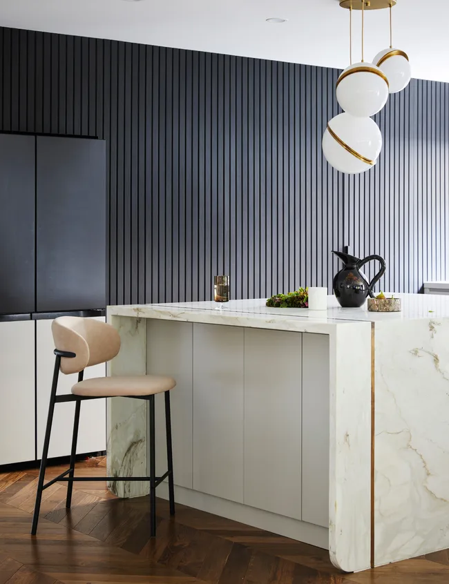 Modern kitchen with marble island, beige chair, dark vertical panel wall, wooden floor, and spherical pendant lights.