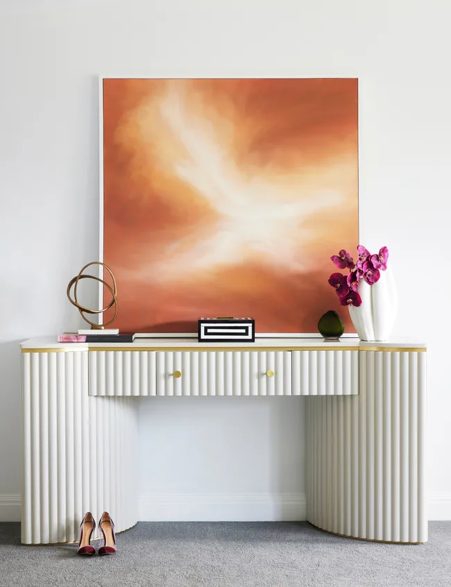 Elegant console table with fluted design, framed abstract art, purple flowers, and neatly placed shoes below on gray carpet.