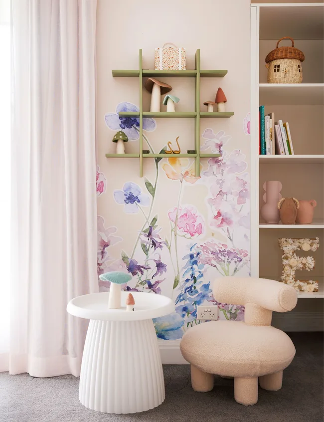 Cozy children's room with floral wall, green shelf, playful decor, round white table, and cream chair.
