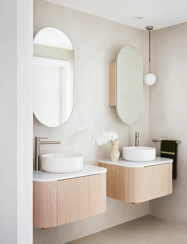 Modern bathroom with two wooden vanities, oval mirrors, and white vessel sinks under pendant light.
