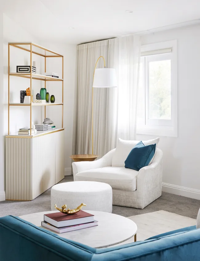 Contemporary living room with white armchair, teal cushion, modern bookshelf, round ottoman, and floor lamp near window.