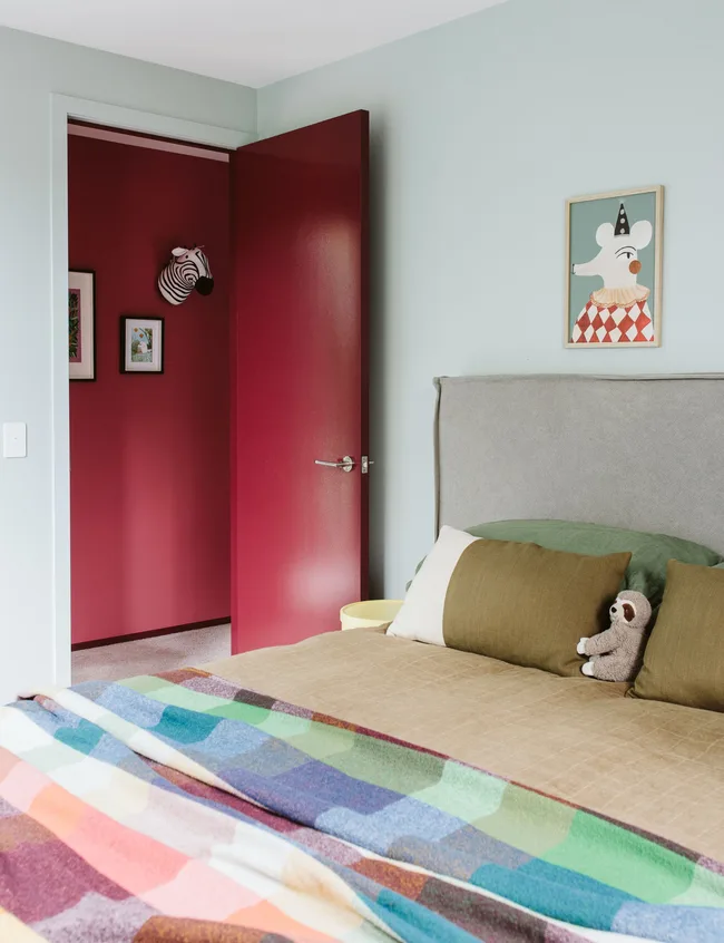 Cozy bedroom with colorful bedspread, stuffed animal, and whimsical wall art. Open red door reveals zebra art in hallway.
