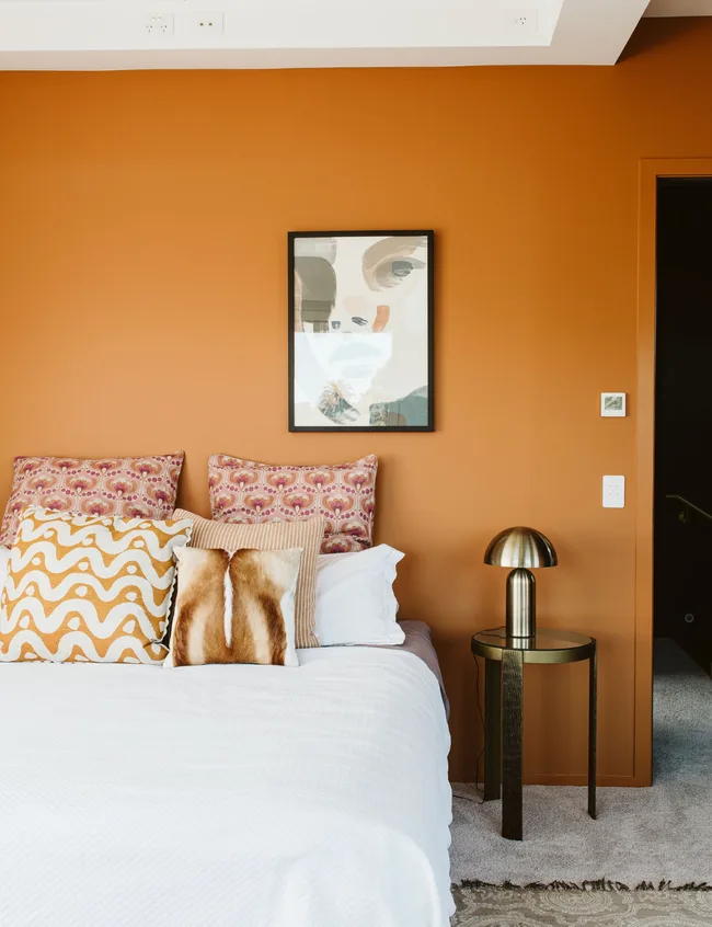 Bedroom with orange walls, abstract art, patterned pillows, white bedding, and a brass lamp on a round side table.