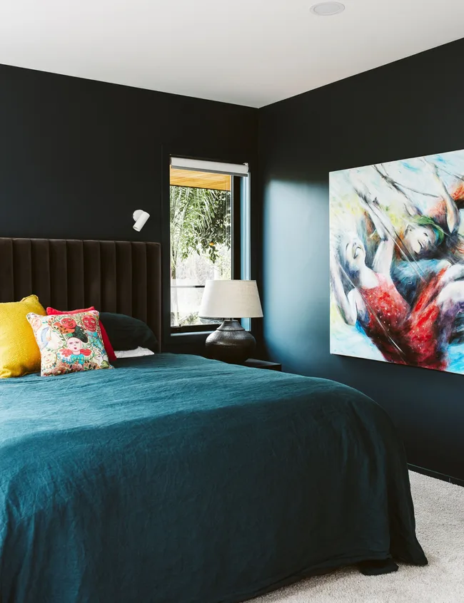 Dark teal bedroom with abstract art, a bed with colorful cushions, and a window showing greenery outside.
