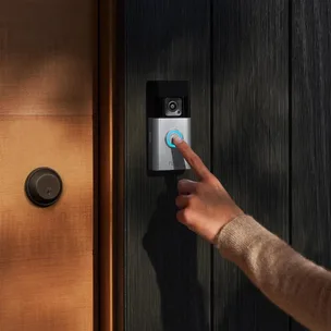 Hand pressing doorbell on modern video doorbell camera mounted next to wooden door with a black finish.
