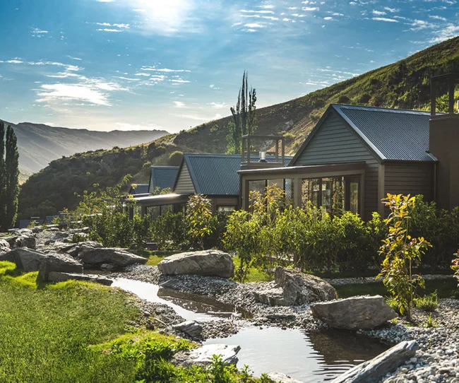 Cabins with metal roofs nestled in a lush mountain landscape beside a small stream on a sunny day.