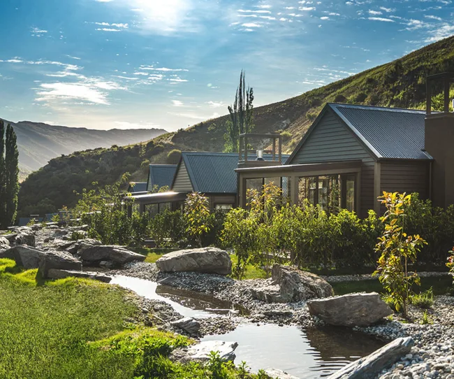 Cabins with metal roofs nestled in a lush mountain landscape beside a small stream on a sunny day.