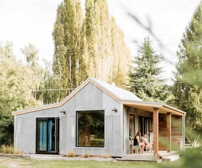 Corrugated metal tiny house with a porch, surrounded by trees, people sitting on the porch with a dog nearby.