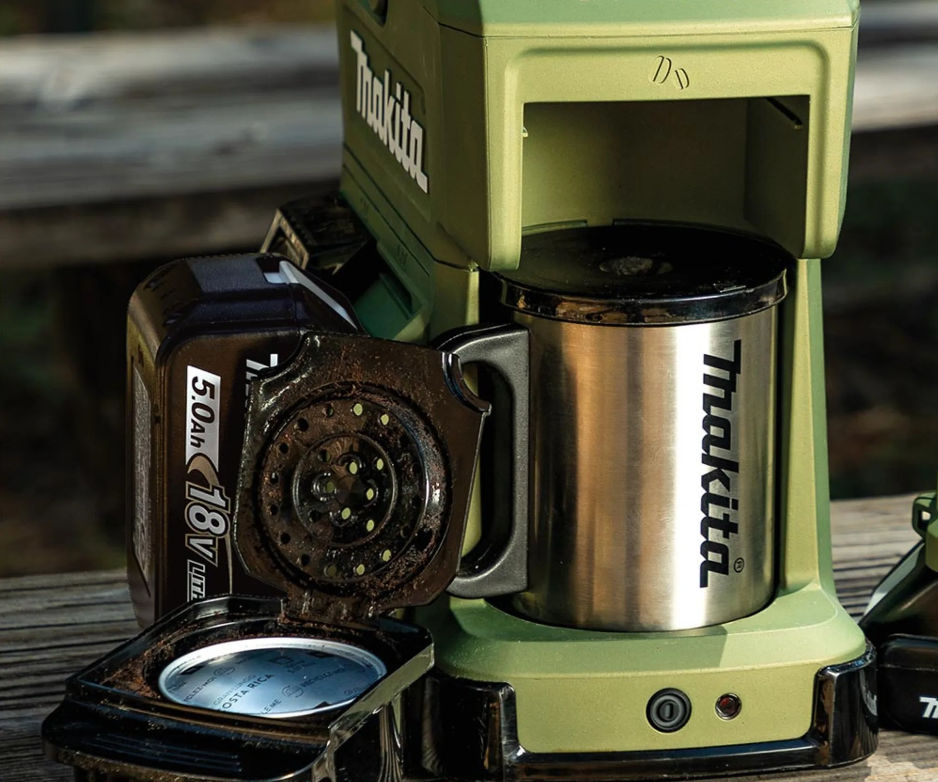 Makita portable coffee maker with stainless steel mug, powered by an 18V battery, on a wooden surface.