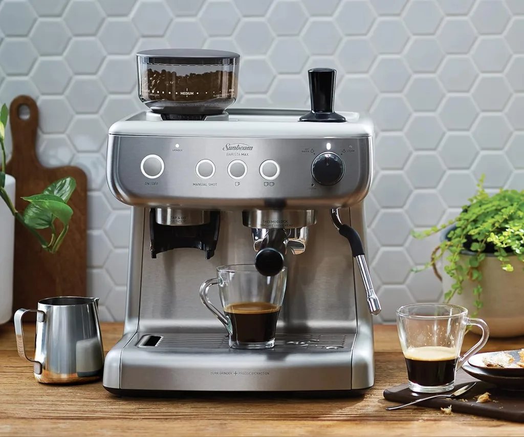 Espresso machine with glass cup of coffee on wooden counter next to milk frother, plant, and cup.